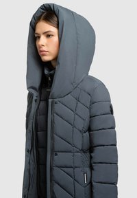 Sivá puffer bunda s veľkou kapucňou, s prešívaným vzorom, so zipsom a elastickými detailmi na bokoch pre priliehavý strih.