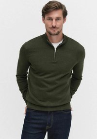 Tumma vihreä, vetoketjuneck pullovari pehmeästä kankaasta, jossa on pitkät hihat ja rento malli. Päällä valkoinen T-paita ja siniset farkut.