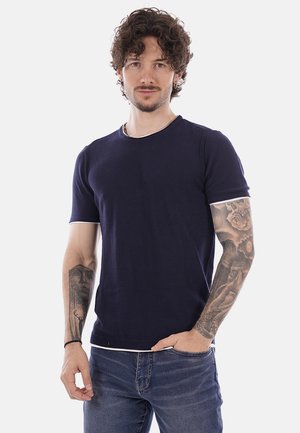 Uomo con capelli ricci, indossa una camicia blu navy a maniche corte e jeans, mostra tatuaggi dettagliati su entrambi gli avambracci, in piedi su sfondo bianco.