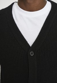 Sort ribbestrikket cardigan med V-hals-design, der har en enkelt sort knap-lukning, båret over en hvid T-shirt.