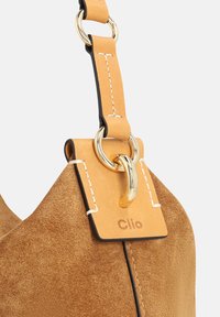 Beigefarbene Wildledertasche mit einem kontrastierenden goldfarbenen Metallring und Lederriemen, versehen mit Ziernähten und einem gravierten Anhänger mit der Aufschrift "Clio".