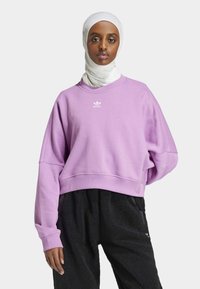 Fialová skrátená mikina s okrúhlym výstrihom a spadnutými ramennými švami; obsahuje biele logo Adidas na hrudi. V kombinácii s štruktúrovanými čiernymi nohavicami.