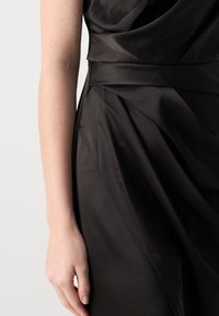 Melna satīna kleita ar drapētu audumu, kurai ir gluda tekstūra un pielāgota vidukļa daļa. Roka novietota uz svārku, izceļot dizainu.