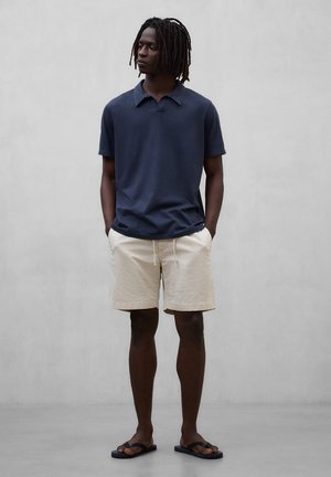 Ung mann med dreadlocks iført en marineblå polotrøye, beige shorts og svarte flip-flops, står foran en ensfarget grå bakgrunn.