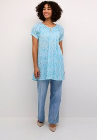 Tunique bleu clair avec un motif floral, manches courtes, détail en trou de serrure au niveau du col, associée à un jean large bleu clair et des chaussures beiges.