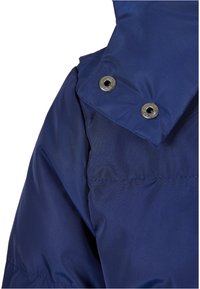 Sezione di giacca imbottita blu navy con colletto alto, dotata di bottoni automatici argentati e una superficie liscia e lucida.
