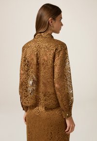 Brown lace blouse featuring long sleeves, a collar, and a semi-translucent design. Decorative floral pattern throughout with button accents.
