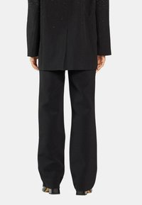 Blazer negro entallado con hombros adornados y mangas largas, combinado con pantalones de pierna ancha a juego y zapatos negros brillantes.