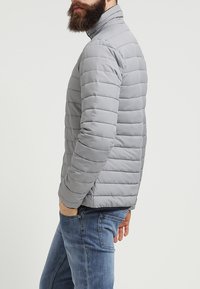 Uomo con barba che indossa una giacca trapuntata grigio chiaro e jeans blu, in piedi con una mano nella tasca, rivolto di lato.