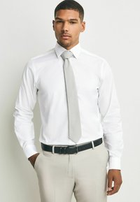 Weißes Hemd mit langen Ärmeln, graue Krawatte und hellbeige Hose. Schwarzer Gürtel mit strukturiertem Finish. Modell steht mit der Hand in der Tasche.