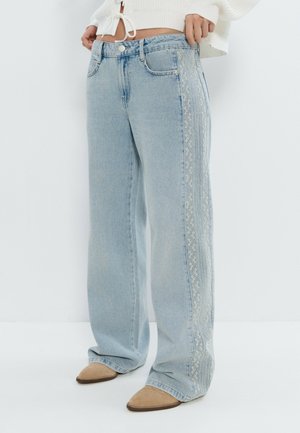 Vaqueros de pierna ancha en azul claro con detalles de encaje a lo largo de las costuras exteriores, combinados con zapatos puntiagudos color cuero y un suéter blanco cropped.