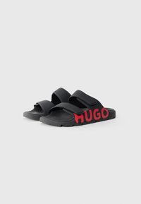 Sandale negre cu talpă plată, dotate cu două curele reglabile, cu tălpi texturate și un branding îndrăzneț "HUGO" de culoare roșie pe lateral. Fabricate din material sintetic.