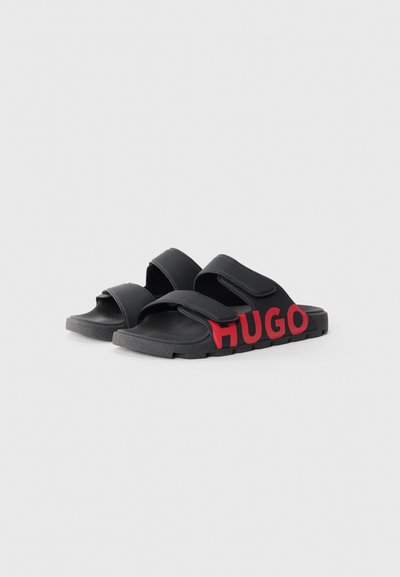 Sandales noires à glissière avec deux sangles réglables, semelles texturées et un logo "HUGO" rouge audacieux sur le côté. Fabriquées en matériau synthétique.