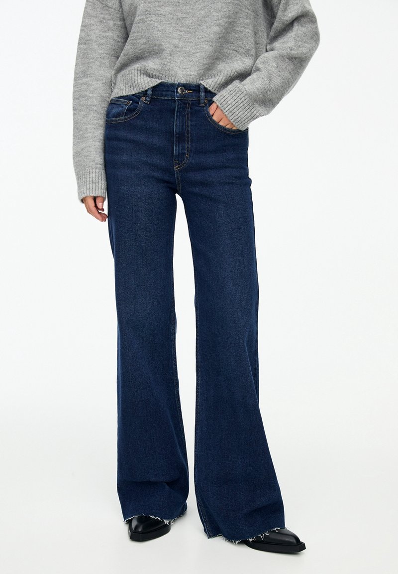 Jeans acampanados de mezclilla azul oscuro con cintura alta, que cuentan con un cierre de botón y cremallera, cinco bolsillos y dobladillos deshilachados. Combinados con un suéter gris.
