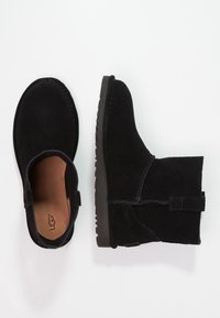 Para czarnych zamszowych botków za kostkę z pętlą do zakupu, płaską gumową podeszwą i tanim wnętrzem, jeden but stoi w pionie, a drugi widok z góry.