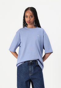 Svetlo modré oversized tričko s krátkymi rukávmi a okrúhlym výstrihom. Na hrudi je malá biela nášivka. Vytvorené s modrými džínsami.