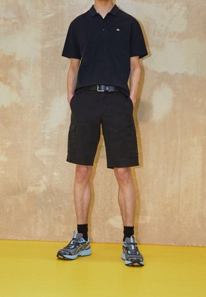 Personne portant un polo noir à manches courtes, un short cargo noir, des chaussettes noires et des chaussures de course bleu et gris, debout sur un sol jaune.
