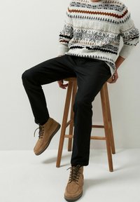 Strickpullover mit weißen, schwarzen und braunen Mustern; schwarze Hosen; braune Wildlederstiefel mit Schnürung; sitzend auf einem Holzschemel.