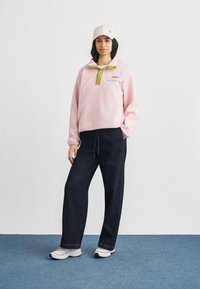 Ροζ φλις πουλόβερ με πράσινη λωρίδα κουμπιών, συνδυασμένο με σκούρο τζιν παντελόνι φαρδύ στα πόδια και ουδέτερα αθλητικά παπούτσια. Άσπρο καπέλο στο κεφάλι.