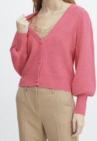 Cárdigan de punto rosa con escote en V, cierre de botones en la parte delantera y mangas voluminosas, combinado con una camiseta de tirantes con encaje debajo.