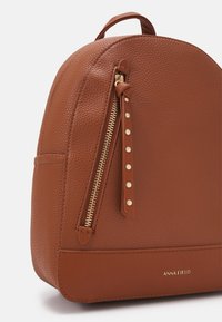 Sac à dos en cuir marron texturé avec une fermeture éclair verticale dorée à l'avant et une languette en cuir cloutée, affichant le nom de la marque "ANNA FIELD" en bas.