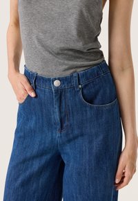 Džínové kalhoty v tmavě modré barvě s rovnými nohavicemi. Mezi funkce patří kapsy, zapínání na stříbrný knoflík a kontrastní stehy.