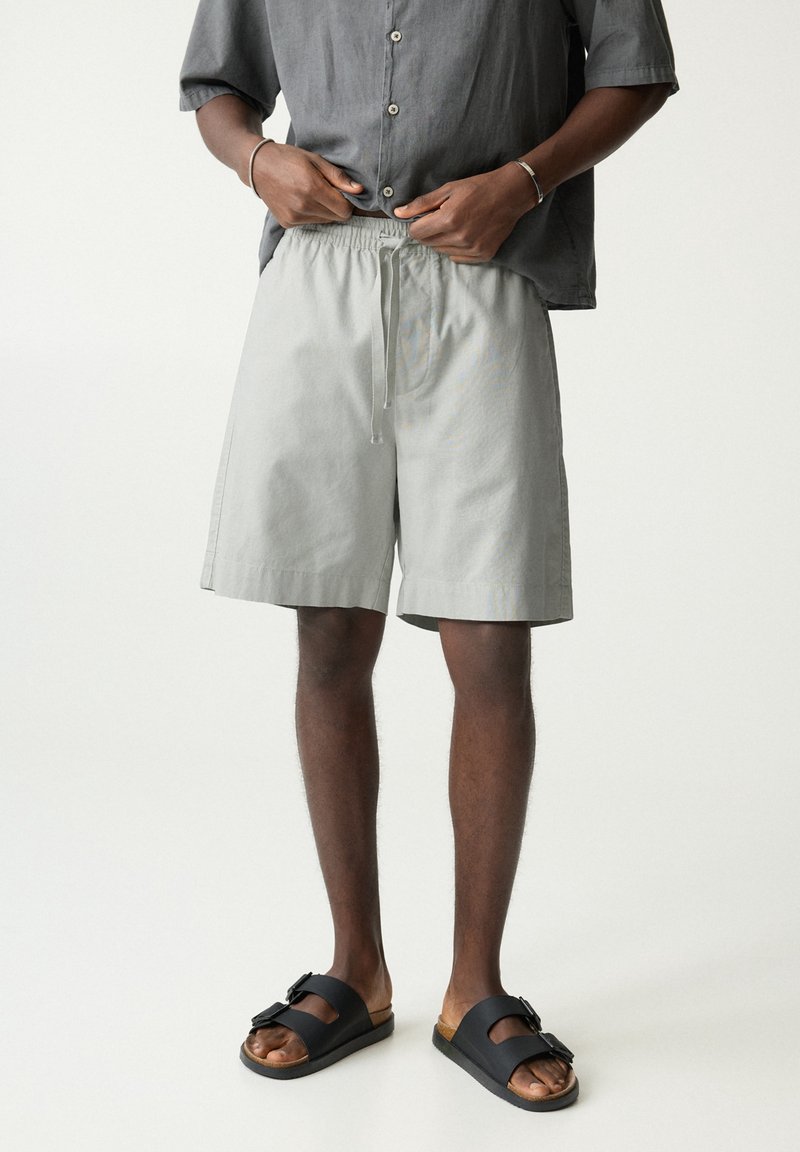 Shorts gris clair avec une taille élastique et un cordon de serrage, offrant une coupe décontractée. Associés à des sandales noires à enfiler.