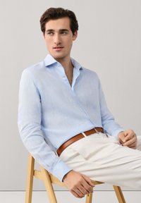 Junger Mann sitzt auf einem Holzstuhl, trägt ein hellblaues Hemd mit Knöpfen, beige Hose und braunen Gürtel, schaut zur Seite.