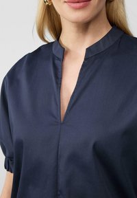Blusa blu navy con colletto alla mandarino e apertura a chiave sulla parte anteriore. Tessuto morbido e liscio con polsini arricciati. Design minimalista.