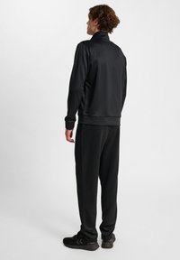 Svart sportig träningsoverall i slät tyg, med hög krage och elastiska ärmslut, tillsammans med stiliga svarta träningsskor.