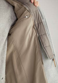 Capa exterior gris con un forro beige suave. Presenta un bolsillo con cremallera y acentos acolchados. Textura suave con un diseño limpio y estructurado.