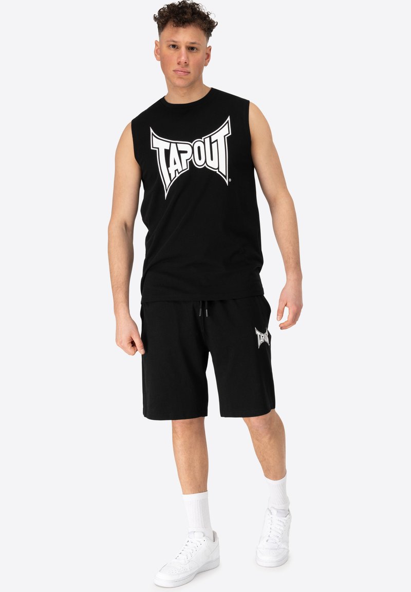 Fekete ujjatlan póló "TAPOUT" logóval, fekete rövidnadrággal, amelyen a logó látható, fehér sportcipővel és fehér bokazoknival párosítva.