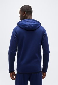 Άνδρας που φοράει σκούρο μπλε φούτερ με κουκούλα και matching παντελόνι, στέκεται γυρισμένος με την πλάτη σε απλό ανοιχτό φόντο.