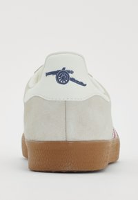 Adidași pentru bărbați cu un corp din suede gri, o etichetă albă pe toc care prezintă un logo al navei de război bleumarin și o talpă din cauciuc texturat de culoare caramel.