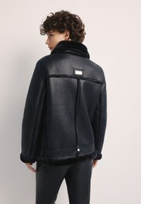 Czarna, skórzana kurtka z teksturowanej skóry z futrzanym kołnierzem i mankietami, z tylną metką i zapięciem napowym na dole.