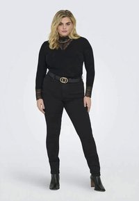 Moteris stovi, vilkinti juodą blizgų viršų su nėrinių detalėmis, juodus džinsus su dvigubos sagties diržu ir juodas aukštakulnes batus prieš šviesią vientisą fono.