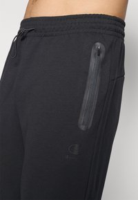 Schwarze Jogginghose mit elastischem Bund, ausgestattet mit einer Reißverschlusstasche und einem kleinen Logo. Hergestellt aus einem weichen, glatten Material.