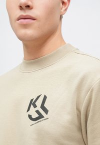 Beige pulóver kerek nyakkivágással, fekete "KJ" logóval és "KARL LAGERFELD JEANS" felirattal a fronton. Puha textúra.