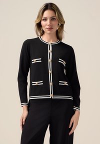 Cardigan nero con bottoni dorati, righe bianche orizzontali su tasche e polsini. Texture liscia, design aderente e forma strutturata.