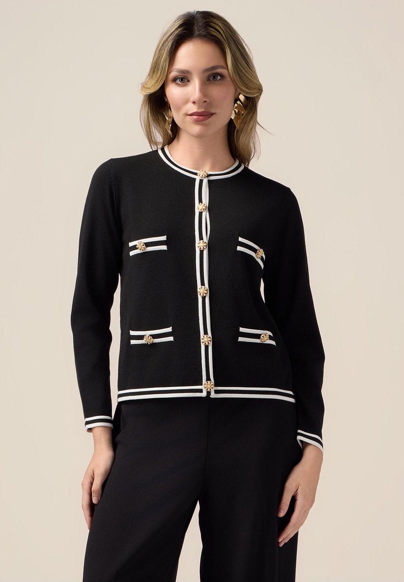 Cardigan nero con bottoni dorati, righe bianche orizzontali su tasche e polsini. Texture liscia, design aderente e forma strutturata.