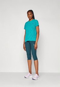 Naine seisab, seljas türkiissinine mustriga lühikeste varrukatega särk, tumedast türkiissinisest lühikesed retuusid ning helesed roosad jooksujalatsid, taustal ühtlane taust.