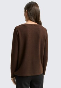 Pull marron avec un tricot texturé, présentant un col large et des manches longues, porté avec un pantalon noir. Détails comprenant des fentes latérales à l'ourlet.