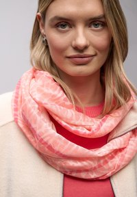 Écharpe légère rose avec un motif texturé et abstrait, portée de manière décontractée. Superposée sur un pull corail, sur un fond neutre.