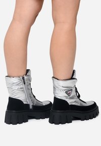 Bottes chelsea argentées avec un design rembourré, accents en suède noir, lacets à l'avant et semelles noires épaisses. Présente un patch logo sur le côté.