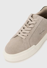 Lys grå semsket sneakers med flate lisser, hvit gummisåle og inngravert Calvin Klein-logo. Har minimalistisk design og glatt tekstur.
