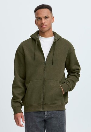 Un giovane uomo si erge con sicurezza indossando una felpa verde oliva con zip sopra una maglietta bianca, con le mani nelle tasche, su uno sfondo chiaro.