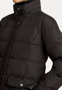 Crna punjena jakna s visokim ovratnikom, s kožnim detaljem na prednjoj džepu i dugmadima na preklop za zatvaranje.