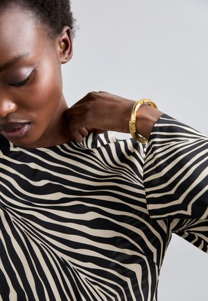 Blouse noire et beige avec un motif de rayures ondulées. Manches longues avec une texture de tissu lisse. Un bracelet en or orne le poignet.