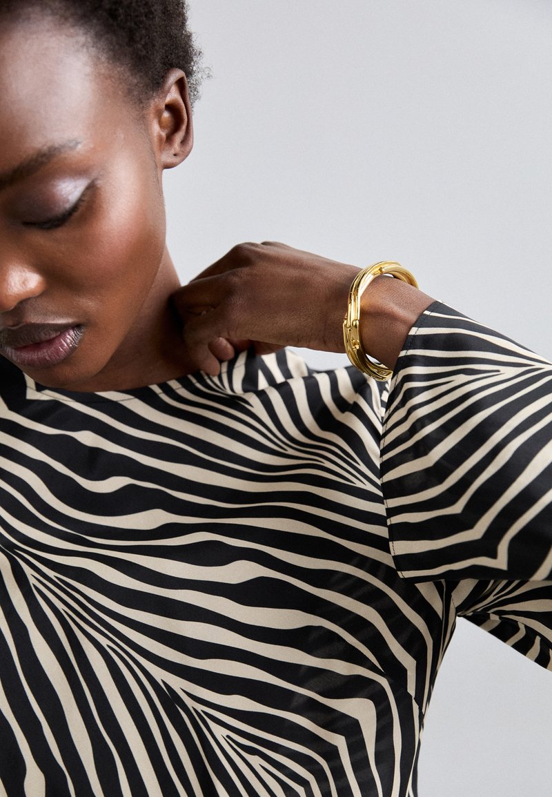 Μαύρη και μπεζ μπλούζα με κυματιστό ριγέ σχέδιο. Μακριά μανίκια με απαλό ύφασμα. Ένα χρυσό βραχιόλι κοσμεί τον καρπό.