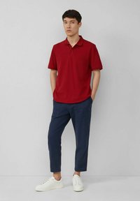 Rotes Polohemd mit klassischem Kragen, kombiniert mit navyfarbener, schmal zulaufender Hose und weißen Sneakers. Schlichtes Design, glatte Textur und einfarbige Farbtöne.